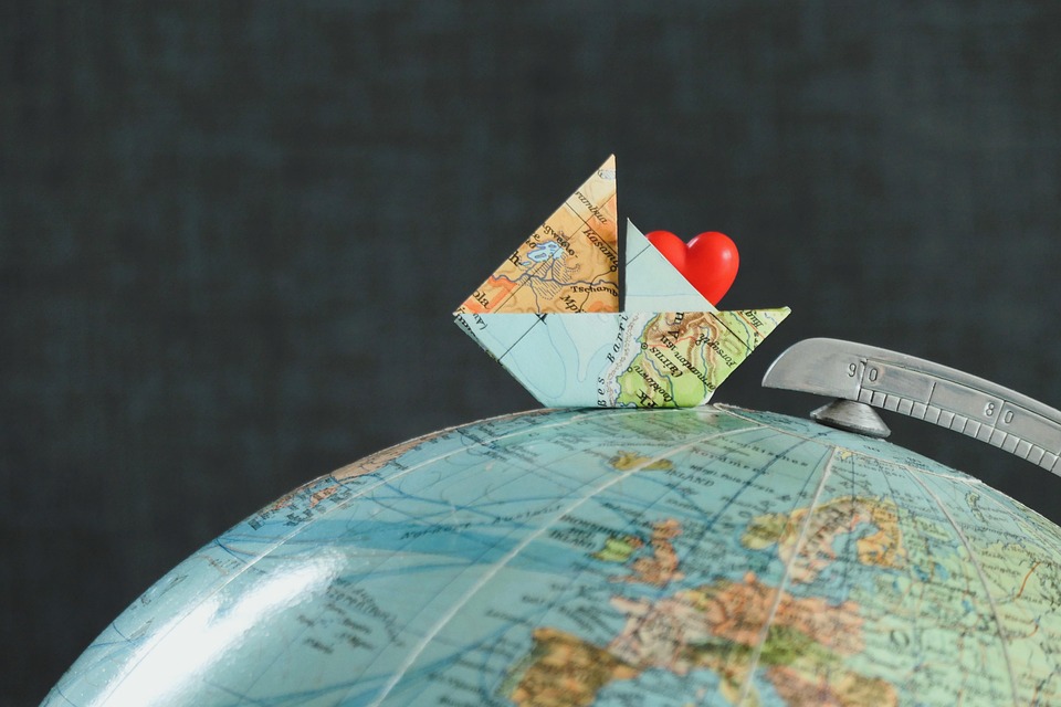 A paper boat made from a world map with a red heart placed on a globe, symbolizing travel and love.